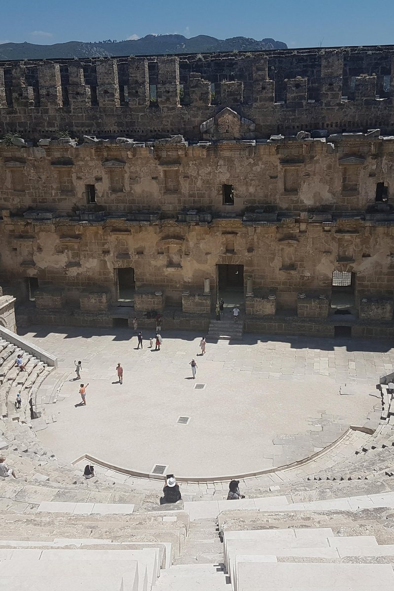 Den höga scenväggen i den romerska teatern i Aspendos reser sig över den svepande halvcirkeln av stenläktare, med besökare som pyttesmå nere i orkestern och Taurus fotberg bortom under en klar himmel. Serik, Antalya, Turkiet.